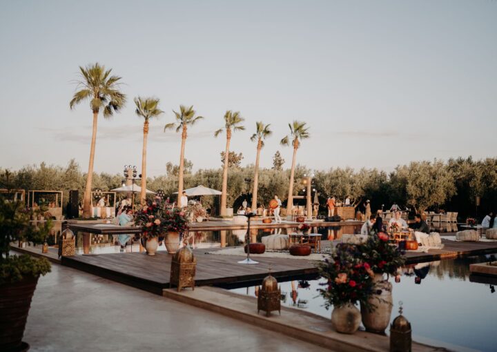 Destination wedding setting in Morocco. There is a wooden deck over a decorative pool, which is surrounded by palm trees and flowers.