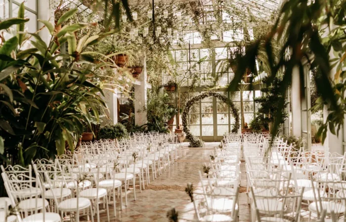 Picture of a large room with rows of white wooden chairs for guests. The place is mostly decorated with green plants.