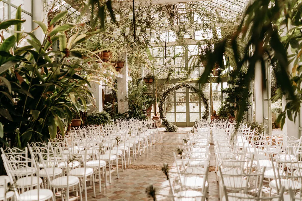 Picture of a large room with rows of white wooden chairs for guests. The place is mostly decorated with green plants.